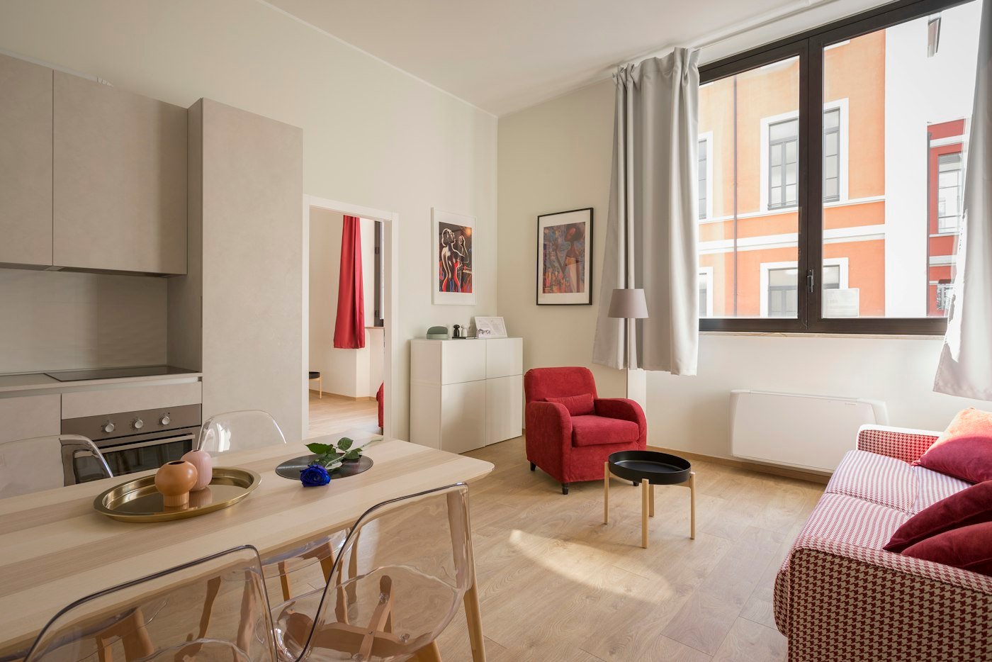 A small flat with a pale wood table, a red armchair, a patterned sofa, and a window onto the street